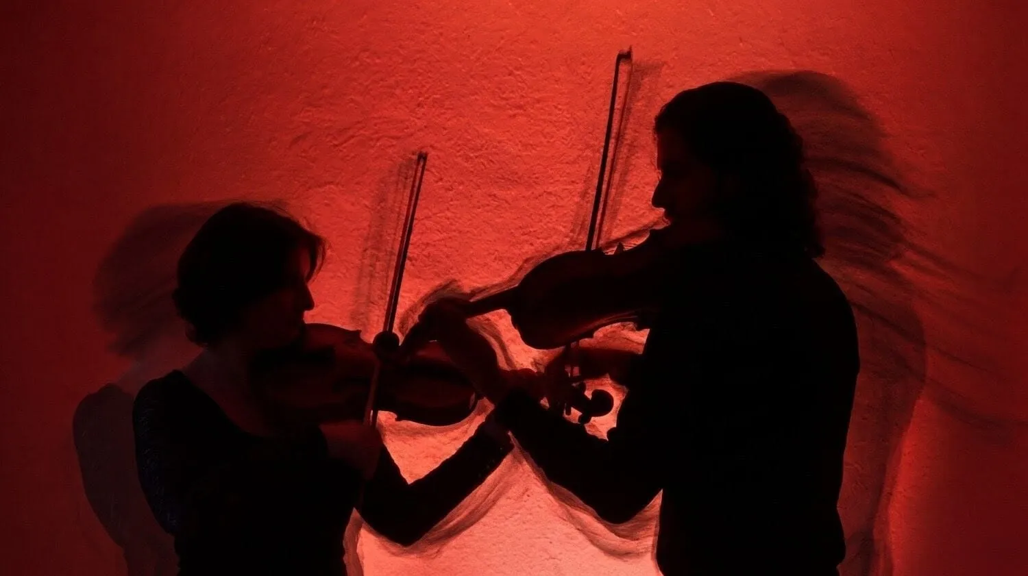 Dos violistas tocan en silueta frente a un muro iluminado en rojo